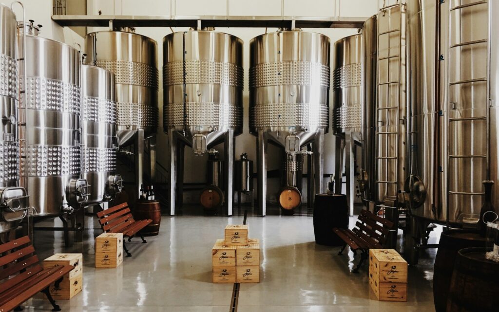 Large stainless steel beer fermentation tanks, wooden crates, and barrels inside a beer production room
