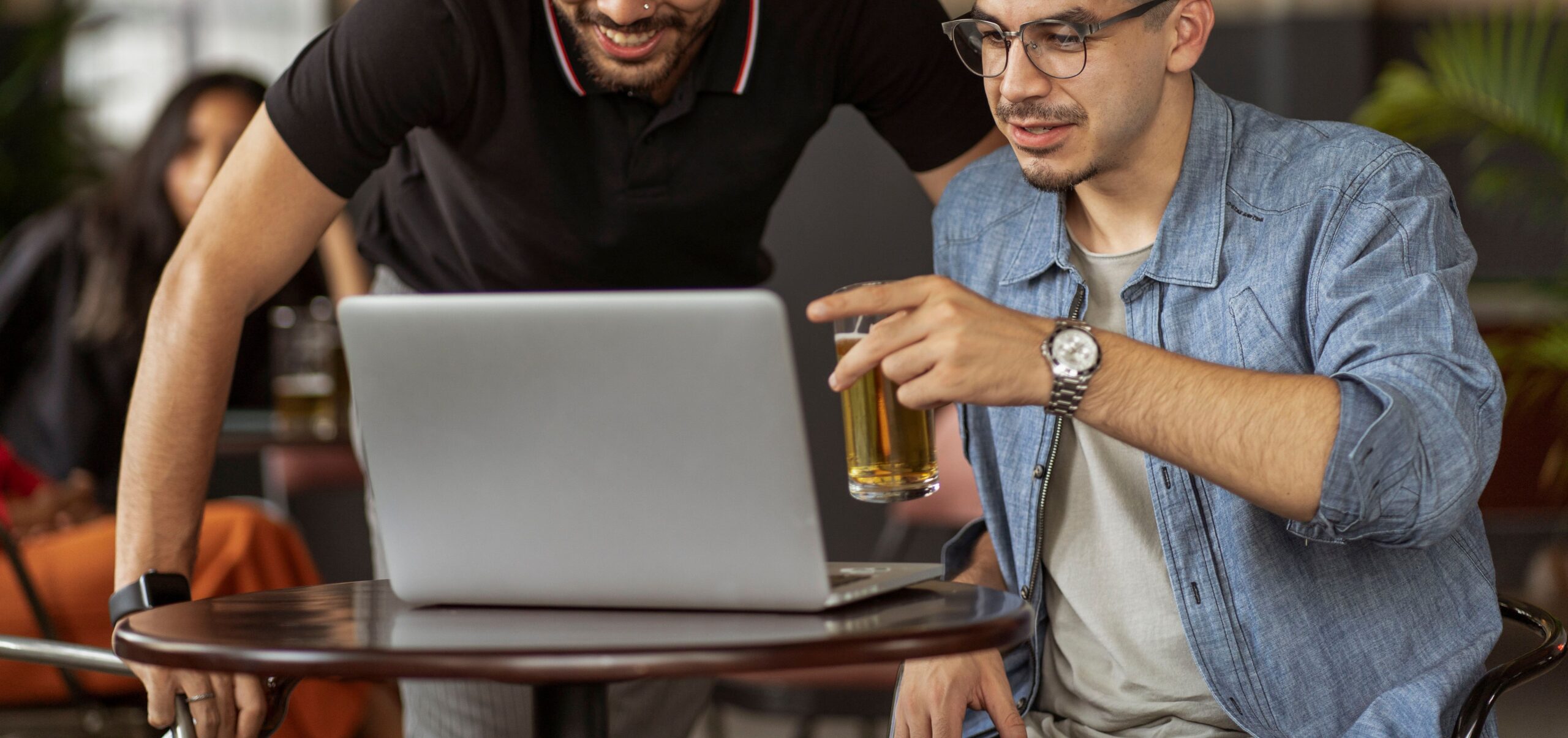 A person holding a glass of craft beer while taking notes on a laptop at a kitchen table